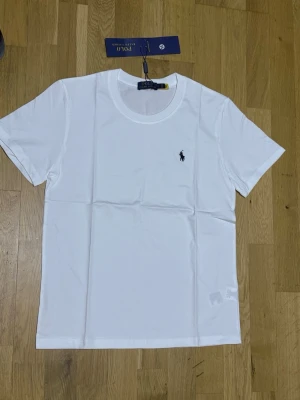 Vit t-shirt från Polo Ralph Lauren - Klassisk vit t-shirt från Polo Ralph Lauren med rund halsringning och liten broderad logga på bröstet. T-shirten är kortärmad och tillverkad i mjuk bomull, perfekt för en clean och stilren look.