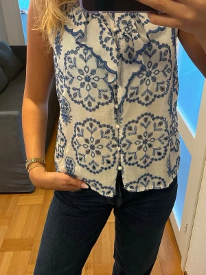 Vit blus med blått broderi och volang - Säljer en ärmlös blus i vitt med blått broderat blommönster och volangdetalj framtill. Blusen har knäppning hela vägen ner och är gjord i ett lätt, luftigt material som känns perfekt för sommaren. Supersnygg till både jeans och kjol!
