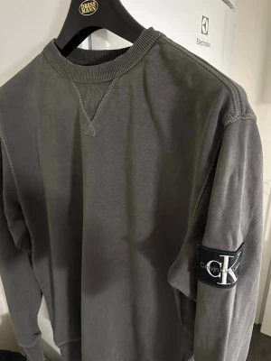 Mörkgrå sweatshirt från Calvin Klein - Nypris 1 295 kr. Endast tvättad två gånger och i mycket fint skick. Snygg mörkgrå sweatshirt från Calvin Klein med rund halsringning och ribbade muddar. Tröjan har en tydlig CK-logga på vänster ärm och dekorativa sömmar vid halsen. Perfekt för en avslappnad och trendig look.