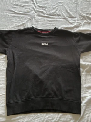 Svart sweatshirt från HUGO BOSS - Snygg svart sweatshirt från HUGO med diskret logga broderad på bröstet. Klassisk rund halsringning, ribbade muddar vid ärmslut och nederkant. Perfekt till jeans eller joggers för en clean och stilren look.