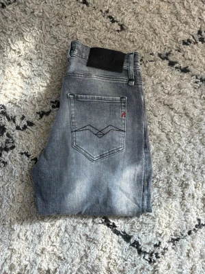 Grå slitna jeans från Replay - Säljer ett par grå jeans från Replay med slitningar och tvättad look. Jeansen har fem fickor, normal midja och raka ben. Snygga detaljer på bakfickan och Replay-logga på linningen. Perfekta för en avslappnad och trendig stil.