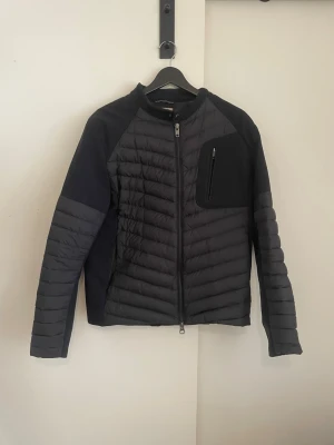 Svart colmar Cardigan - Snygg svart pufferjacka med quiltad design och dragkedja framtill. Jackan har en praktisk bröstficka med dragkedja och kontrasterande tyg på ärmar och sidor. Perfekt för kalla vinterdagar och har en modern, sportig look.    Den har lite slitningar men det är smått och syns inte så mycket därav de låga priset om ni vill se de är de bara att slå en pling! Som sagt de märks inte! Size 50 står det skulle gissa att det är runt M passar mig som är 180 72 kg.