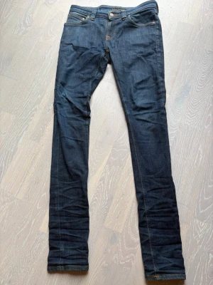 Nudie jeans i färgen blå - Snygga mörkblå jeans från märket nudie med raka ben och klassisk femficksmodell. Jeansen har kontrastsömmar i gult och dekorativa broderier på bakfickorna. Tillverkade i slitstarkt denimtyg som ger en tidlös look. Perfekta till en avslappnad stil.