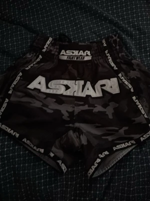 Svarta kamouflage fight shorts Askari - Säljer ett par helt nya svarta fight shorts från Askari med grått kamouflagemönster och tydlig logga på framsidan. Shortsen har elastisk midja och är perfekta för kampsport eller träning. Materialet är lätt och snabbtorkande.