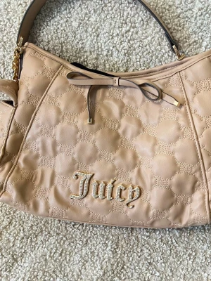Beige handväska från Juicy Couture - Snygg beige handväska från Juicy Couture med quiltat mönster och guldfärgade detaljer. Väskan har ett dekorativt band framtill och ett axelband med kedjedetalj. Insidan är fodrad med logotypmönster i svart på ljus bakgrund. Använd bara en gång.