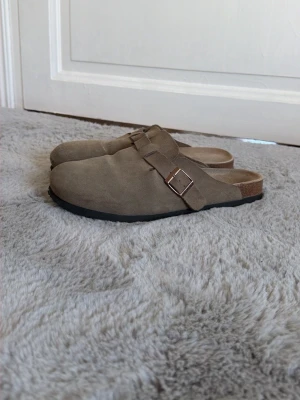 Beige mockatofflor från Birkenstock - Säljer ett par klassiska beige tofflor från Birkenstock i mocka med justerbart spänne i metall. De har en bekväm fotbädd och svart sula, perfekta för chill dagar hemma eller när du vill ha det extra bekvämt.