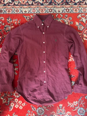 Vinröd skjorta från Ralph Lauren - Klassisk vinröd skjorta från Ralph Lauren i slim fit-modell. Skjortan har långa ärmar, knappar framtill och button-down-krage. Tillverkad i mjuk bomull och har den ikoniska loggan broderad på bröstet.