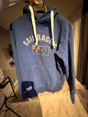 Vintage Sail Racing hoodie  - Blå hoodie från Sail Racing med vit dragsko i huvan och stor ficka framtill. På bröstet står det 'Sail Racing' och en flagga är tryckt under texten. På ena ärmen finns siffran 77 i rött och vitt. Perfekt för en sportig och avslappnad stil.