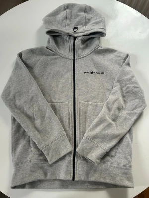 Grå hoodie med dragkedja Sail Racing - Snygg grå hoodie från Sail Racing med svart dragkedja framtill och två stora fickor. Tröjan har huva med broderad logga och diskret Sail Racing-tryck på bröstet. Tillverkad i mjukt sweatshirtmaterial, perfekt för chill dagar.