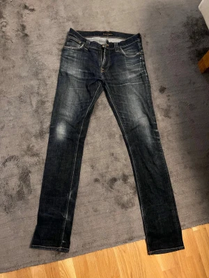 Nudie slim fit jeans - Säljer ett par mörkblå nudie slim fit jeans med snygga slitningar och kontrastsömmar. Jeansen har fem fickor, normal midja och klassisk knappgylf. Materialet är denim i bomull med lite stretch för extra komfort. Köpt på Åhléns i sthlm för 3 månader sen, knappt använda.
