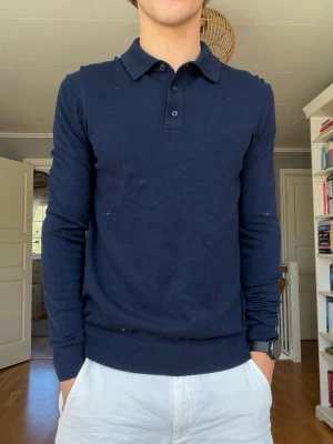 Mörkblå långärmad pikétröja – stilren slim fit - Stilren mörkblå långärmad pikétröja med klassisk krage och tre knappar framtill. Snygg, relativt kroppsnära passform och ribbad mudd både vid ärmslut och nederkant.  Fint begagnat skick med normalt lätt bruksslitage. Väldigt lättmatchad modell som fungerar bra både till vardags och mer uppklätt.