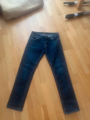 Nudie Jeans Tight Long John blå jeans storlek 28/30 - Snygga mörkblå jeans med rak passform och klassisk femficksdesign. Jeansen har kontrastsömmar och detaljerade bakfickor med broderi. Perfekta för en avslappnad och trendig look. Materialet är denim i bomull. Sytt ner de så storleken är 28/30 ungefär, osäker på storleken.