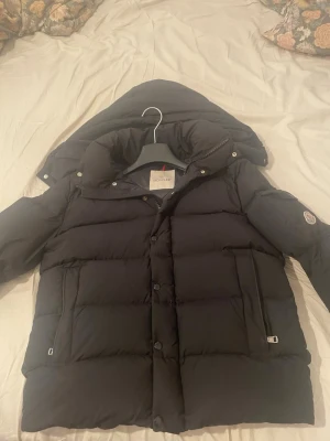 Moncler verzere  - Säljer en svart dunjacka från Moncler med avtagbar huva och klassisk Moncler-logga på ärmen. Jackan har stora, vadderade paneler, dragkedja och knappar framtill samt två fickor med dragkedja. Perfekt för kalla vinterdagar.