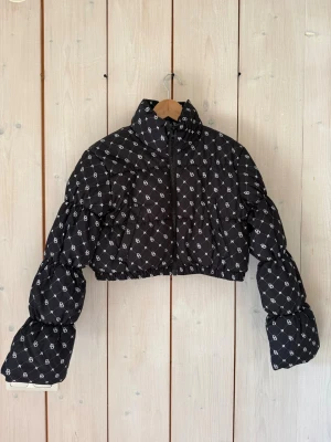 Svart cropped pufferjacka med mönster - Snygg svart cropped pufferjacka med vitt monogrammönster över hela jackan. Jackan har hög krage, dragkedja framtill och puffiga ärmar med resår vid ärmslut och midja. Nyskick. Aldrig använd.