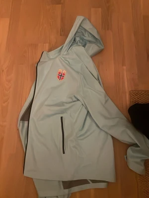 Ljusblå Nike vindjacka med huva - Säljer en ljusblå vindjacka från Nike med huva och Storm-FIT-material. Jackan har ett norskt märke på bröstet och svarta dragkedjor på fickorna. Perfekt för blåsiga dagar och sportiga outfits.