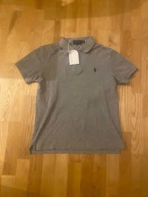 Grå pikétröja från Polo Ralph Lauren - Klassisk grå pikétröja från Polo Ralph Lauren med korta ärmar och krage.  Det är storlek M men passar storlek S