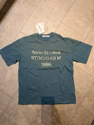 Blå Acne Studios t-shirt med tryck - Säljer en blå t-shirt från Acne Studios med vit text framtill där det står 'Acne Studios STOCKHOLM 1996'. Klassisk passform och rund hals. Perfekt för dig som gillar stilrena plagg med en twist.