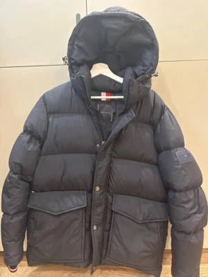 Svart dunjacka från Tommy Hilfiger - Säljer en svart dunjacka från Tommy Hilfiger med stor huva och två stora fickor framtill. Jackan har dragkedja och tryckknappar, samt diskret logga på ärmen. Perfekt för kalla vinterdagar och riktigt snygg streetstil.