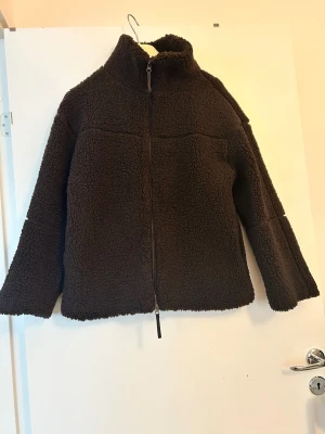 Mörkbrun teddyjacka med dragkedja - Mysig mörkbrun teddyjacka med hög krage och hel dragkedja framtill. Jackan har en boxig passform och är gjord i fluffigt teddy-material som håller dig varm. Perfekt för kyliga dagar och enkel att styla till olika outfits.