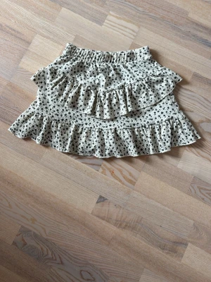 Blommig volangkjol från Zara - Söt kjol från Zara i storlek 164 med beige botten och små svarta blommor. Kjolen har flera volanger och elastisk midja som ger en skön och trendig passform. Perfekt för dig som gillar romantiska detaljer och en lekfull stil. Skort