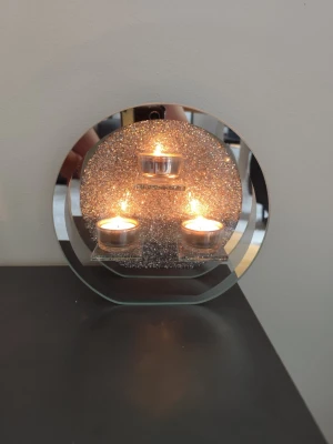 Rund glittrig ljushållare i glas - Säljer en rund ljushållare i glas och metall med glittrig bakgrund och plats för tre värmeljus. Modern design med spegelkant och små dekorstenar som ger extra bling. Perfekt som dekoration i vardagsrummet eller sovrummet.