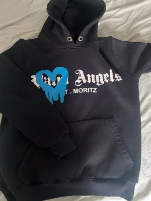 Svart hoodie med blått hjärta - Säljer en svart hoodie med stor huva och magficka. På bröstet står det 'Palm Angels St. Moritz' i vitt och ett blått, droppande hjärta tryckt ovanpå. Hoodien har ribbade muddar och är gjord i mjukt sweatshirtmaterial.
