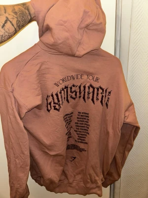 Gymshark  - Säljer en hoodie från Gymshark med stor svart text och tryck på ryggen samt mindre logga framtill. Hoodien har huva och en stor magficka. Tillverkad i mjukt bomullsmaterial och har ribbade muddar vid ärmslut och nederkant. Perfekt för en avslappnad stil. Är xs herr men funkar till tjej mes