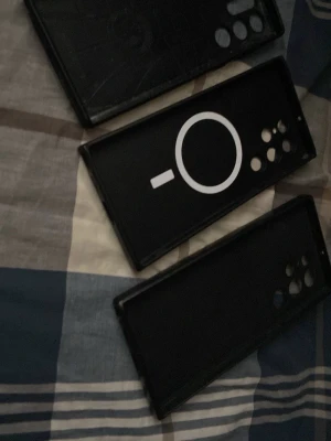 Samsung Galaxy S24 Ultra mobil skal  - Tre svarta Samsung Galaxy S24 Ultra mobilskal i plast eller silikon med flera kamerahål, ett av skalen har en vit MagSafe-liknande cirkel på insidan. Passar större smartphones med flera kameralinser. Stilrena och skyddande skal som ger ett enkelt och modernt intryck.
