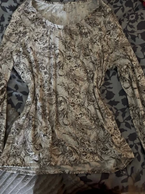 Mönstrad långärmad topp i beige - Säljer en beige långärmad topp med detaljerat paisleymönster i svart och brunt. Toppen har rund halsringning och är tillverkad i ett mjukt, stretchigt material som sitter snyggt på kroppen. Perfekt för dig som gillar unika prints och vill sticka ut lite extra.