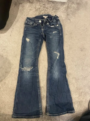 Blå bootcut jeans från Gina Tricot - Säljer ett par blå bootcut jeans från Gina Tricot i storlek 152. Jeansen har slitna detaljer, utsvängda ben och klassisk femficksmodell. Snygg tvätt med kontrastsömmar och trendiga slitningar på låren och knäna.
