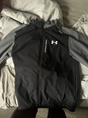 Svart och grå vindjacka Under Armour - Snygg svart och grå vindjacka från Under Armour med dragkedja framtill, bröstficka med logga och huva. Jackan har sportig design och är tillverkad i ett lätt, vindtätt material som passar perfekt för utomhusaktiviteter.