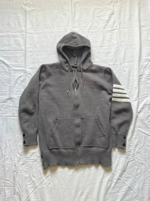 Grå hoodie med segelbåtsmotiv - Säljer en grå hoodie med dragkedja och huva. På vänster ärm finns tre vita ränder och på ryggen ett stort segelbåtsmotiv i vitt, svart och rött. Hoodien har fickor framtill och knappar vid ärmslut och nederkant. Perfekt för dig som gillar unik design. Vet inte exakt märket men köpte den för mycket. Pris går att diskuteras då tröjan är lite nopprig men inget som inte går att lösa enkelt.