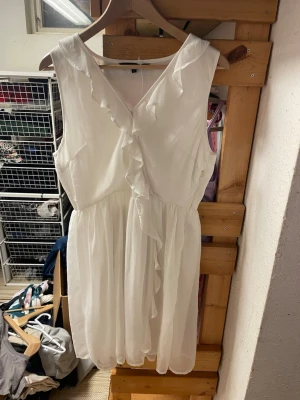 Vit ärmlös klänning med volang - Söt vit klänning med v-ringning och volangdetaljer framtill. Klänningen är ärmlös och har en luftig, kort modell med markerad midja. Perfekt för varma dagar och enkel att styla med accessoarer. Tillverkad i ett lätt och skönt material.