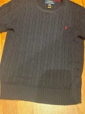 Mörkblå kabelstickad tröja Polo Ralph Lauren - Den är 152 cm lång jätte fin Kabelstickad mörkblå tröja från Polo Ralph Lauren med rund halsringning och den klassiska röda logotypen broderad på bröstet. Tröjan har ribbade muddar vid ärmslut och nederkant, perfekt för en stilren look.kommer med lapp 