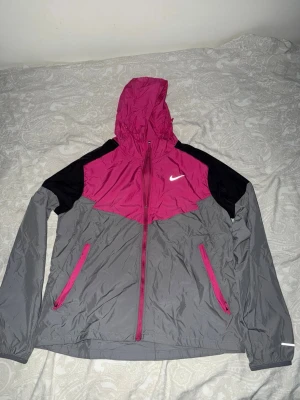 Nike miler - Snygg vindjacka från Nike i grått, svart och starkt rosa. Jackan har huva, dragkedja framtill och två fickor med rosa dragkedjor. Materialet är tunt och vindtätt, perfekt för blåsiga dagar. Ikonisk Nike-logga på bröstet och sportig design.