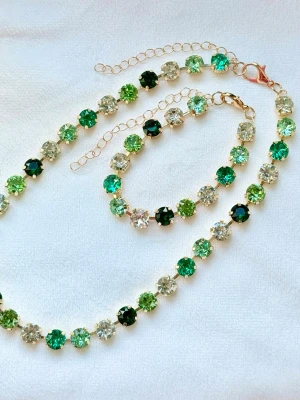 Grönt smyckeset  - Snyggt halsband och armband  med runda 8 mm glittriga stenar i olika gröna och klara nyanser. Stenarna sitter i guldfärgade fattningar och har en justerbar kedja med karbinlås. Perfekt för dig som vill ha något färgglatt och glittrigt till din outfit. 