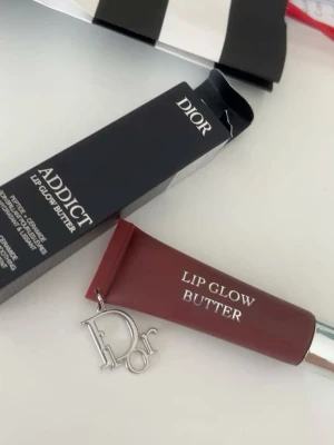 Dior Addict Lip Glow Butter - Lyxigt läppbalsam från Dior i tub med charmig Dior-berlock. Färgen är en djup bärig nyans och formulan är krämig och återfuktande. Förpackningen är elegant med silverdetaljer och tydlig logga. Perfekt för dig som vill ha mjuka och glansiga läppar.