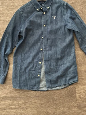 Blå långärmad skjorta från Gant - Snygg blå skjorta från Gant i bomull med klassisk button-down krage och broderad logga på bröstet. Skjortan har vita knappar och en stilren, tidlös design som passar perfekt till jeans eller chinos.
