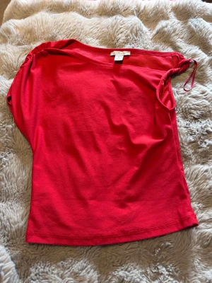 Röd topp med snörning på axeln - Snygg röd topp med snörning på ena axeln som ger en unik touch. Toppen är ärmlös och har en enkel, rak passform. Tillverkad i mjukt bomullstyg som känns skönt mot huden. Perfekt för dig som vill ha en färgstark och stilren look.