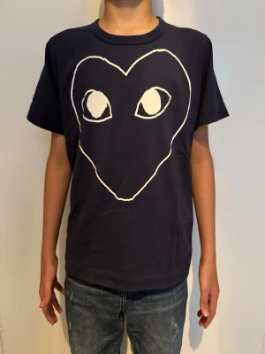 Mörkblå t-shirt med hjärtmotiv - Snygg mörkblå t-shirt med ett stort vitt hjärta och ögon tryckt på framsidan. T-shirten har rund halsringning och korta ärmar. 