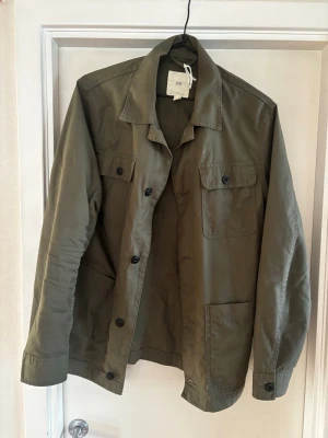 Olivgrön overshirt från H&M - Stilren olivgrön overshirt från H&M med klassisk krage, fyra fickor framtill och svarta knappar. Tillverkad i ett slitstarkt bomullstyg och har en rak passform. Perfekt lager på lager-plagg. Hör av er vid frågor, priset går att diskutera 💯