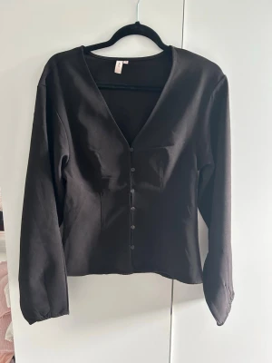 Svart långärmad blus med knappar - Stilren svart blus med lång ärm och djup v-ringning. Blusen har knäppning framtill med små svarta knappar och är figursydd för en snygg passform. 