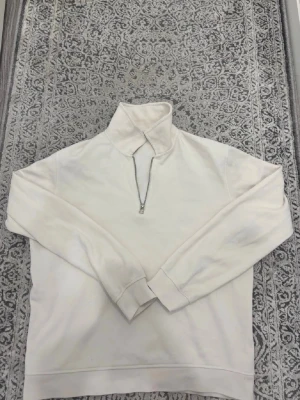 Vit halfzip hoodie - Säljer en stilren vit hoodie med halfzip och hög krage. Tröjan har långa ärmar, muddar vid ärmslut och nederkant samt en mjuk bomullskänsla. Perfekt för dig som gillar en clean och enkel look.