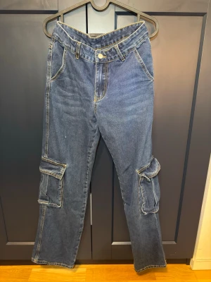 Blå cargobyxor i denim - Snygga blå cargobyxor i klassisk denim med raka ben och stora fickor på sidorna. Byxorna har kontrastsömmar, bälteshällor och knappgylf. Perfekta för en avslappnad och trendig streetwear-look.