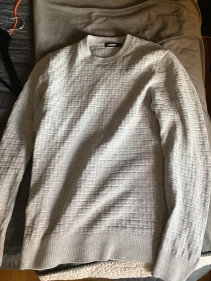Ljusgrå tröja från J.Lindeberg - Snygg ljusgrå tröja från J.Lindeberg med diskret rutmönster i strukturen. Tröjan har rund halsringning, ribbade muddar och är tillverkad i ett mjukt material som känns skönt mot huden. Perfekt för dig som gillar stilrena plagg.