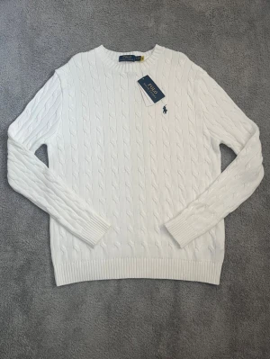 Vit kabelstickad tröja Polo Ralph Lauren - Vit kabelstickad tröja från Polo Ralph Lauren med rund halsringning och klassisk svart logga broderad på bröstet. Tröjan har ribbade muddar och är tillverkad i mjukt material som ger en clean och stilren look. Har bara testats men har ej använts i vardagen. Det finns ett litet ursprungligt fel på den tredje bilden men som ej är synligt om man använder den.