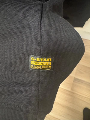 Svart sweatshirt från G-Star RAW - Snygg svart sweatshirt från G-Star RAW med rund halsringning och diskret logga på bröstet. Tröjan har en gul patch med G-Star Originals RAW Denim på sidan och ribbade muddar vid ärmslut och nederkant. Perfekt till jeans eller cargo.