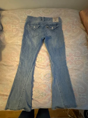 Ljusblå bootcut jeans från true religon  - Säljer ett par ljusblå bootcut jeans från true religon  med klassiska fickor bak och fram. Jeansen har utsvängda ben och detaljerade sömmar. Perfekta för dig som gillar retrostil och vill ha ett par bekväma jeans med snygg passform. Storlek 29, pris kan diskuteras 