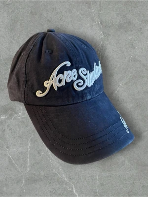 Mörkblå keps från Acne Studios - Snygg mörkblå keps från Acne Studios med broderad logga i vitt framtill och på skärmen. Klassisk böjd skärm och justerbar rem bak. Tillverkad i bomullstyg som ger en avslappnad och trendig look.