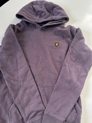 Lila hoodie med gul logga - Säljer en lila hoodie från lyle scoot  med huva och magficka. På bröstet finns en liten gul logga i form av en fågel. Hoodien har ribbade muddar vid ärmslut och nederkant. Perfekt för en avslappnad och cool stil.